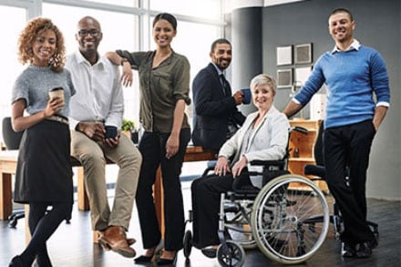 six people standing and sitting in front of a desk four women one in wheelchair and two men