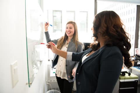 Marketing: Two women talking in front of a white board while writing in a office with window in background